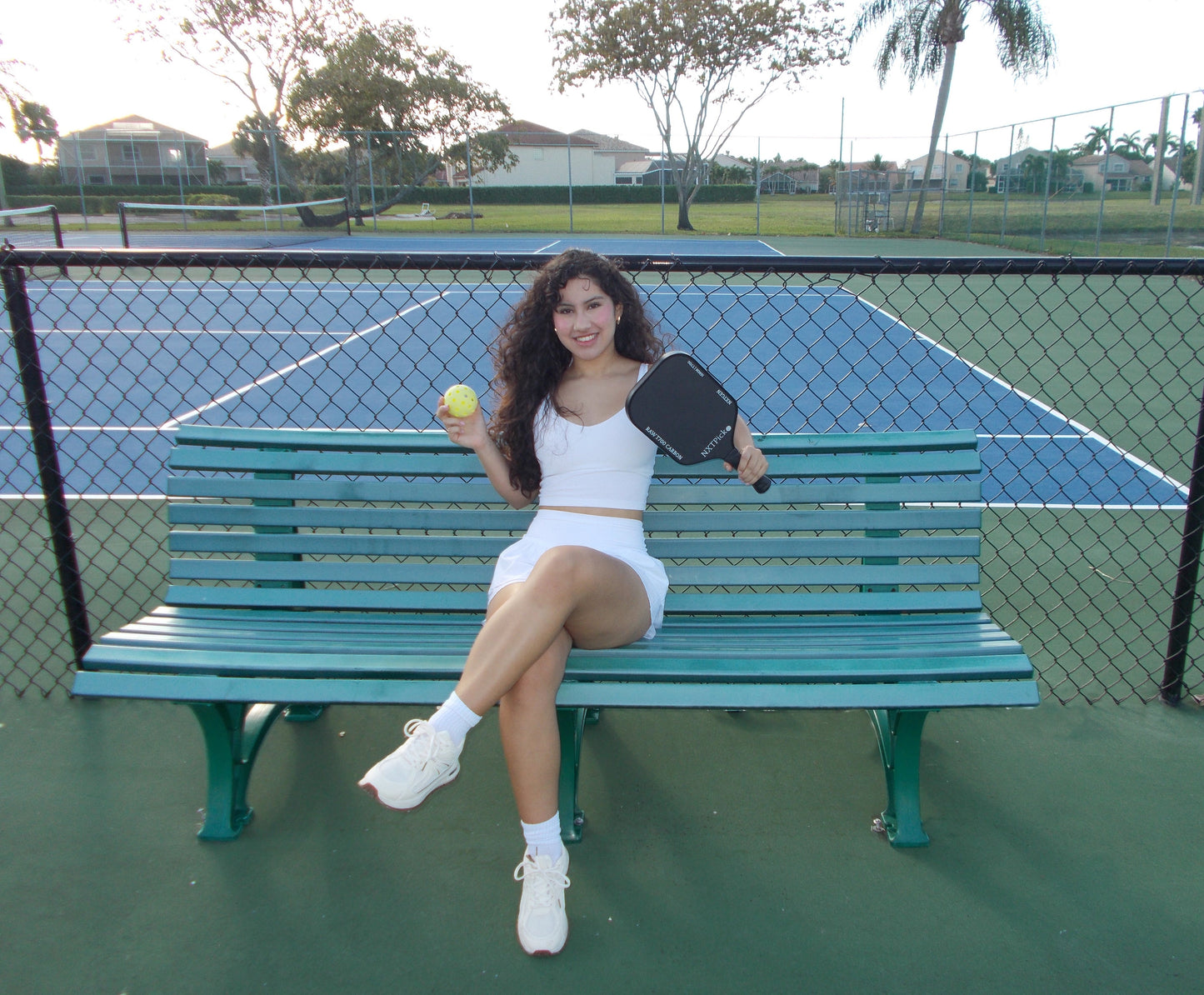 Woman sitting on tennis bench holding a NXTPick carbon fiber pickleball paddle and ball on court