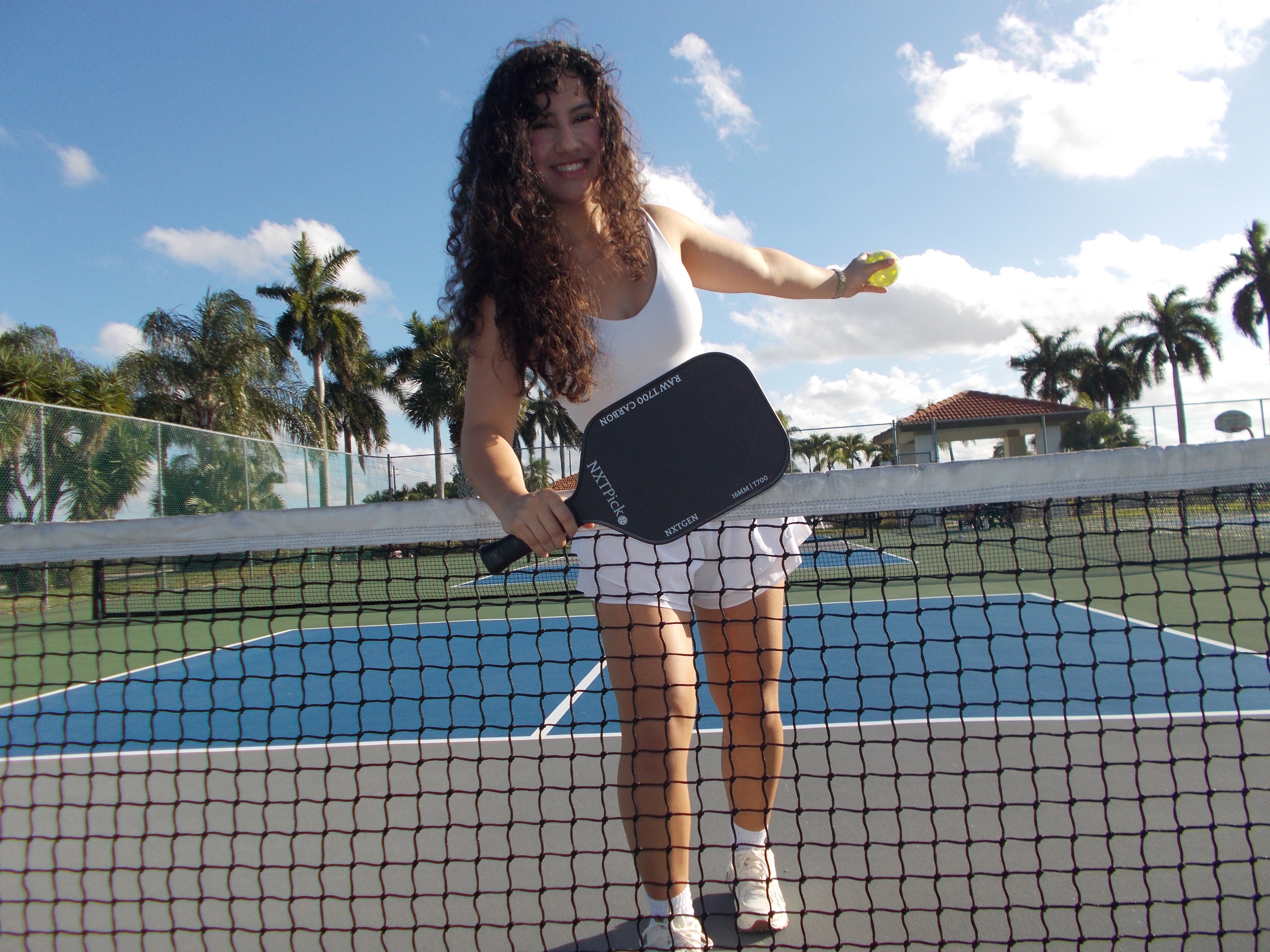 Woman smiling and holding a NXTPick carbon fiber pickleball paddle at the net on outdoor court